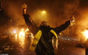 A yellow-vested protester raises his arms with cars burning behind him in scenes of violent carnage in Paris last night