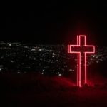 red neon cross on top of hill w city lights