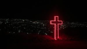 red neon cross on top of hill w city lights