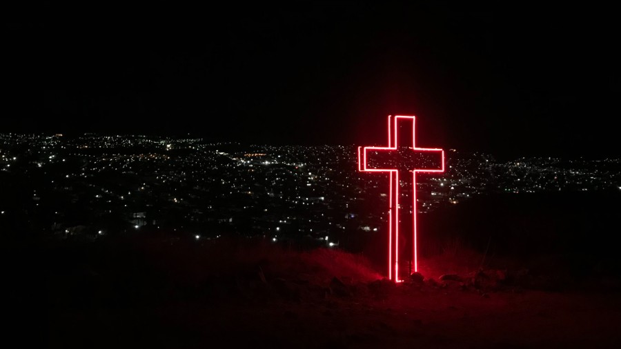 red neon cross on top of hill w city lights