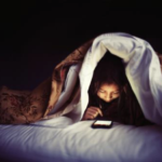 Child reading mobile device under blanket at night