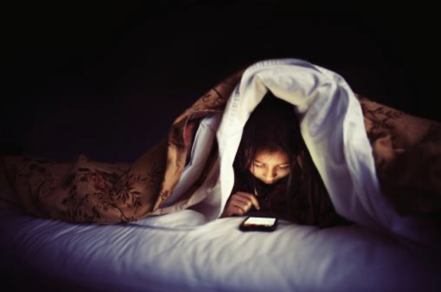 Child reading mobile device under blanket at night
