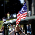 man waves US Flag from car window during parade